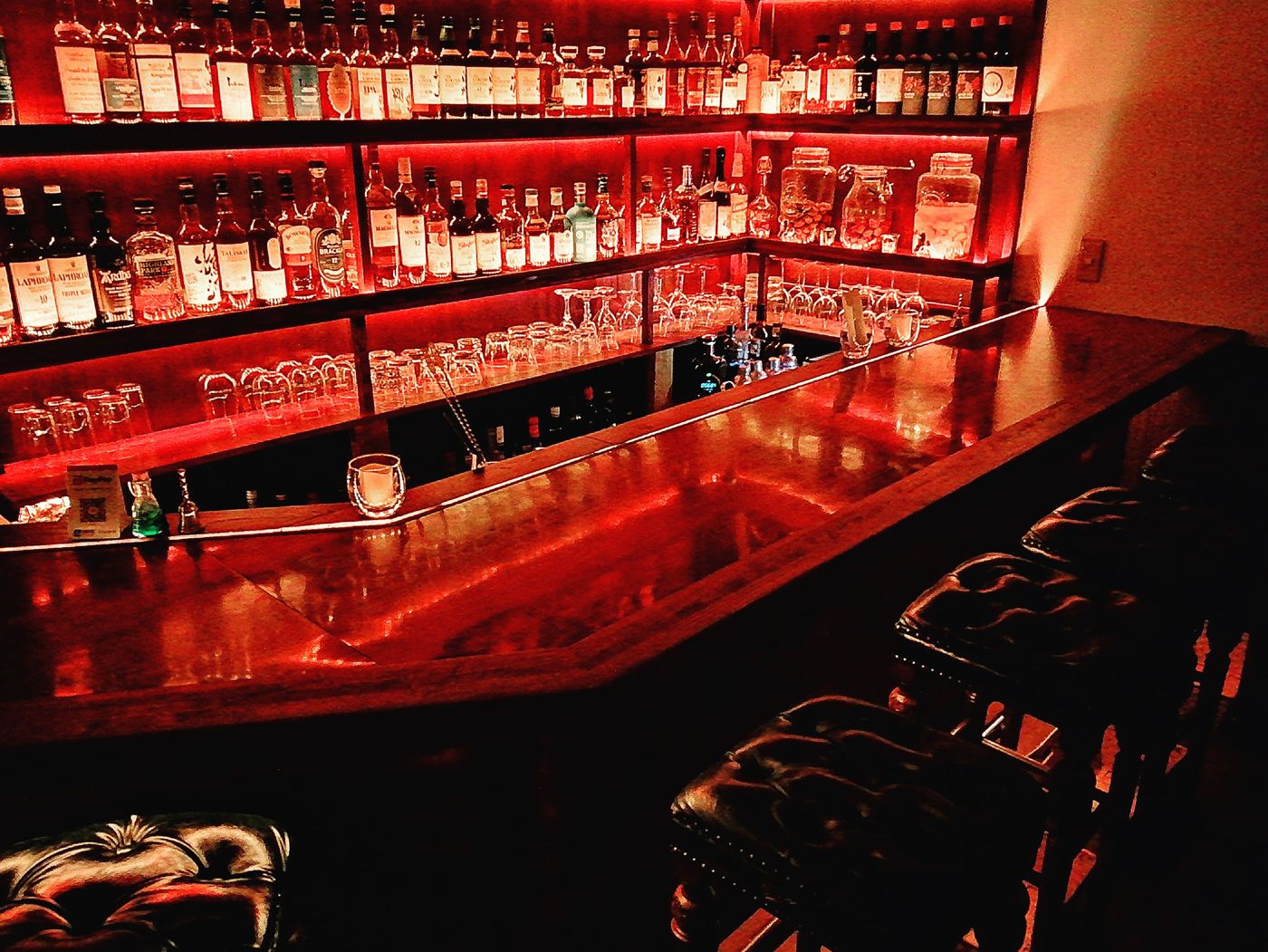 A long wooden bar counter with leather stools, glowing red under shelves of whisky
