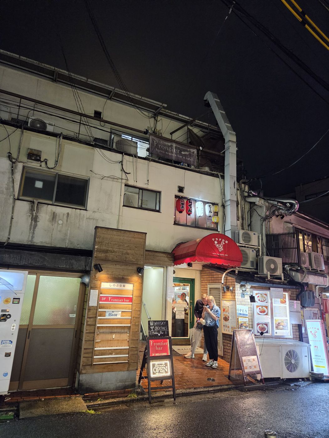The Shinobu Kaikan building at night, with a red Frontier bar sign and a Whisky Cocktails Hidden Bar board at the entrance